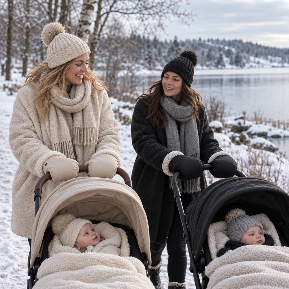 Zwei Mamas mit Kinderwagen im Schnee – eine mit beigen, eine mit schwarzen Lovelle Cosy Kinderwagen-Handwärmern
